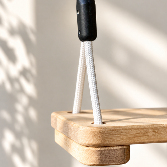 A close-up of a wooden climbing wall or playground component with a white rope and a black plastic connector, set against a warm beige wall with soft leaf shadows.