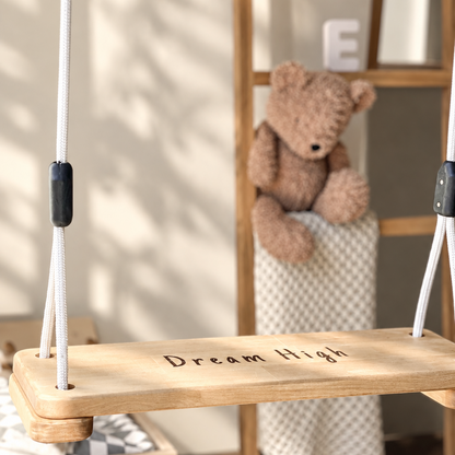 A wooden swing seat with the words 'Dream High' printed on it, hanging by white cords in a softly lit room. A brown teddy bear sits on a knitted blanket in the background against a wooden shelf.