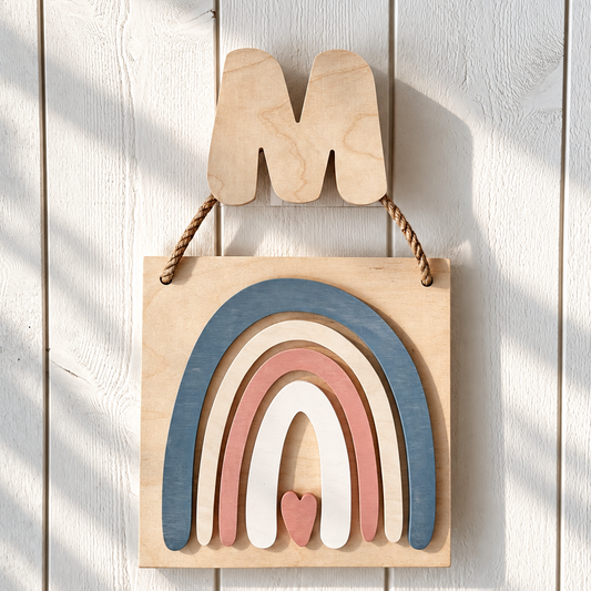 A wooden hanging wall decor piece shaped like the letter M above a layered rainbow design in blue, pink, cream, and natural wood tones, displayed against a white wood plank wall with soft shadow overlays.