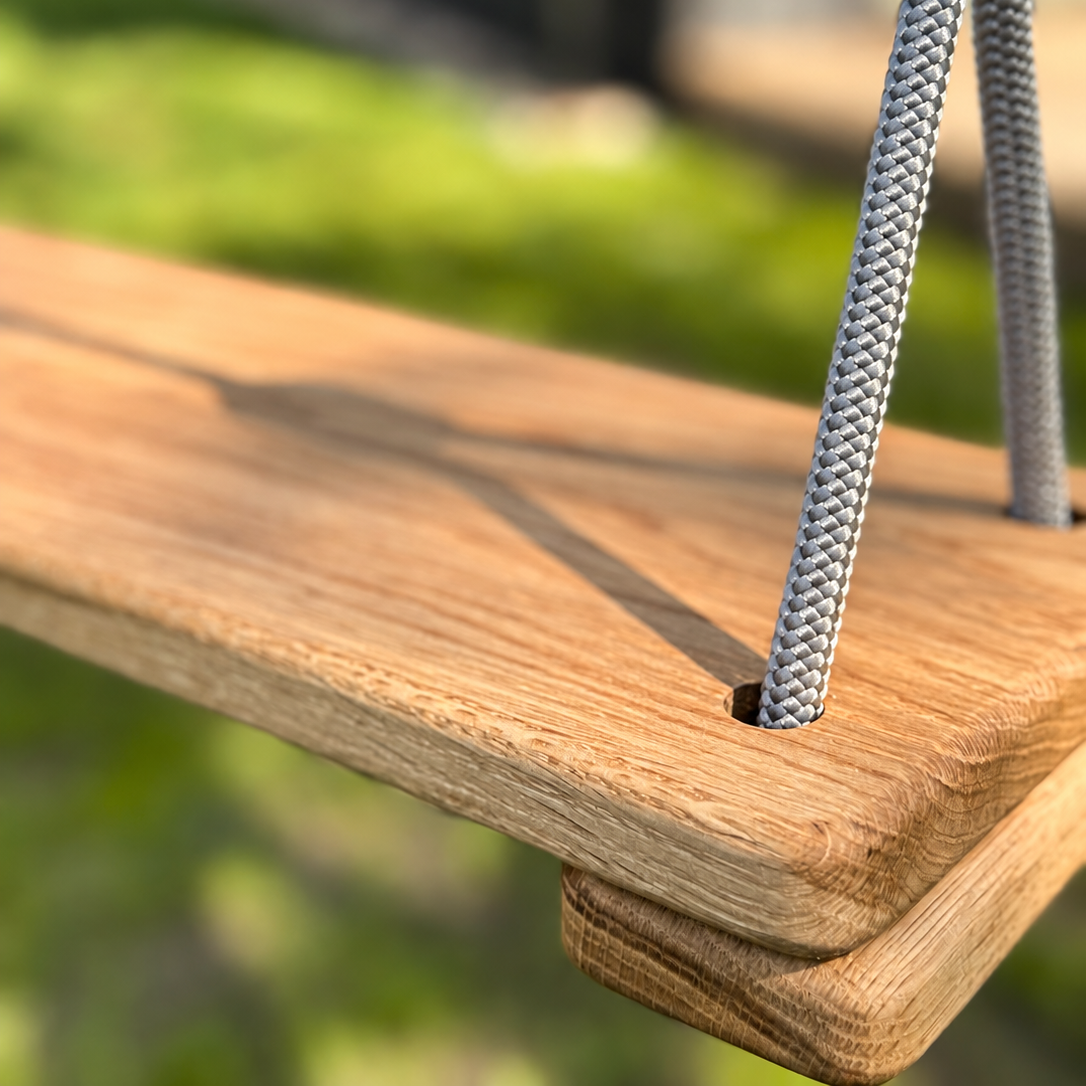 Close-up of a wooden swing seat with thick gray braided hanging ropes and a black plastic top, photographed outdoors with a blurred green garden background.