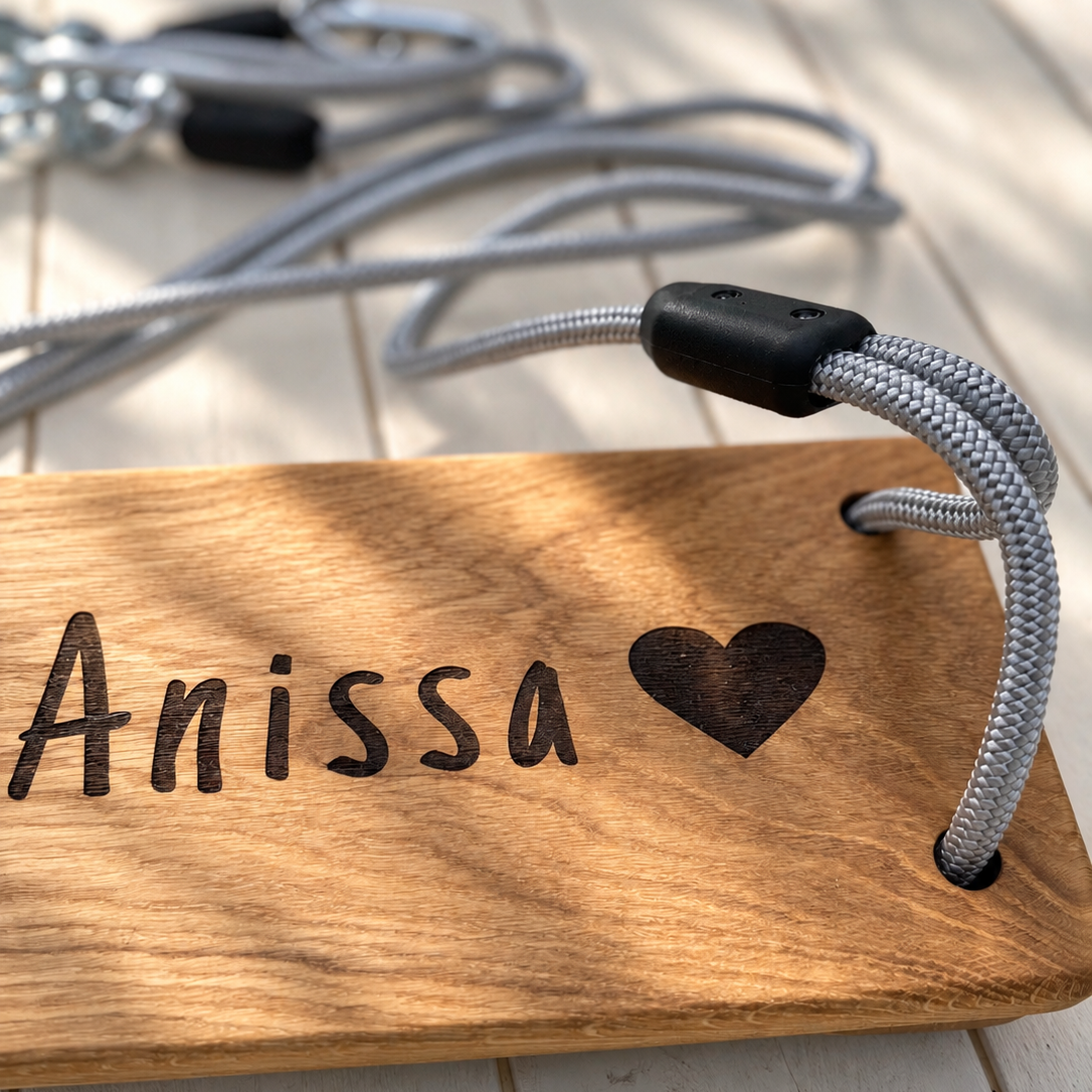 A close-up of a personalized wooden tag engraved with the name 'Anissa' and a heart, attached to a gray braided rope with a black toggle, resting on a light wood plank surface.