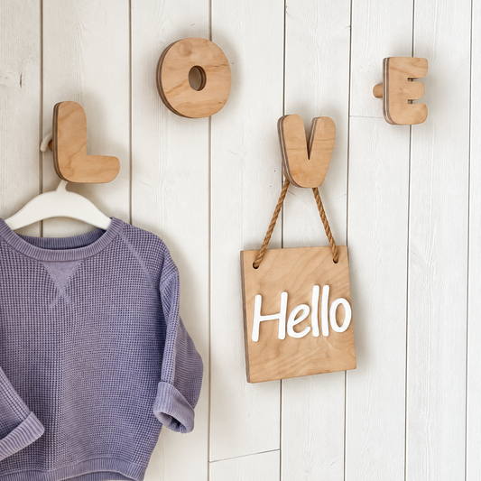 Wooden wall hooks spelling LOVE hanging on a white wood-paneled wall, with a lavender long-sleeve shirt on a hanger and a small wooden sign that says Hello.